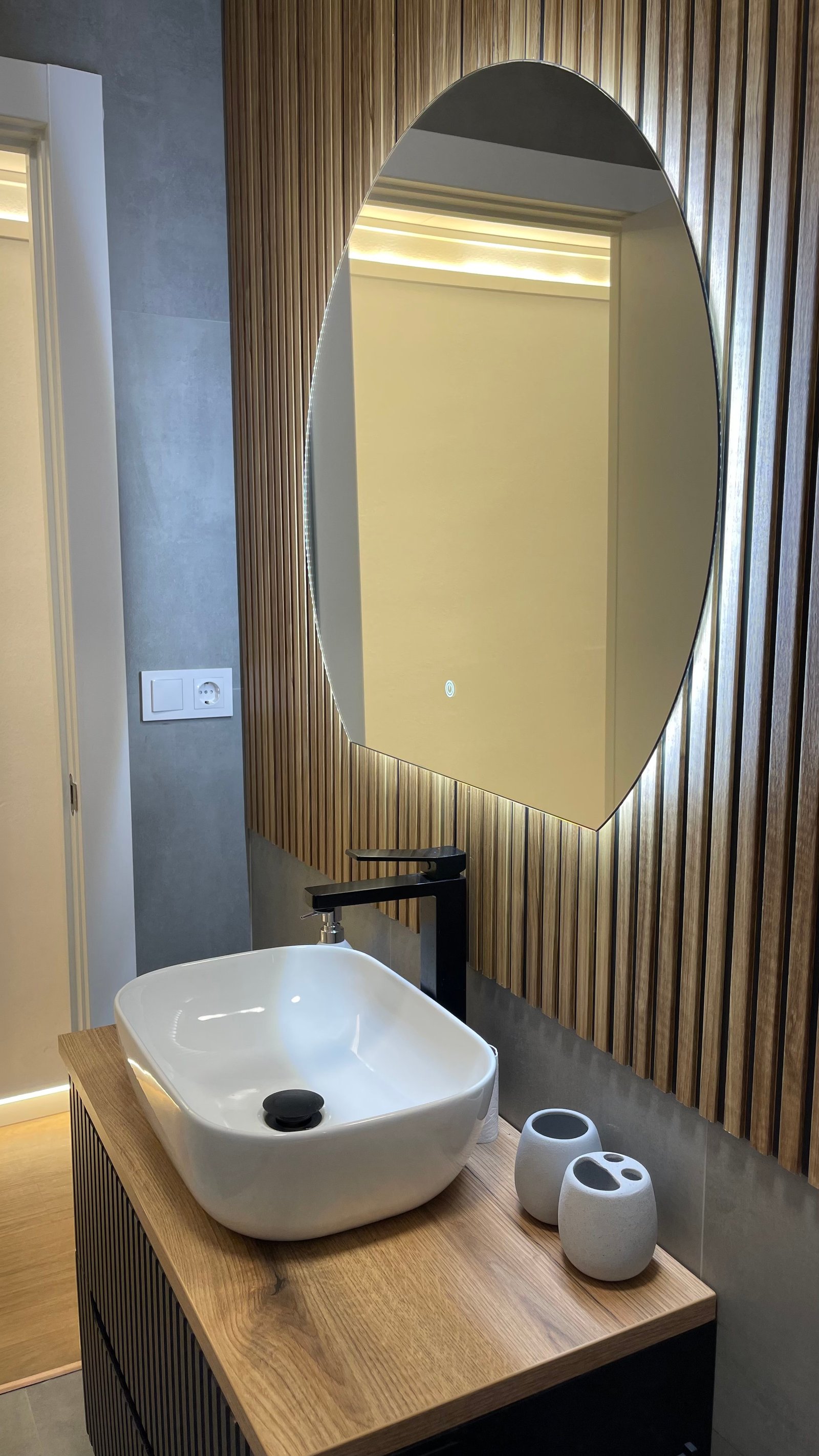 Baño con espejo ovalado retroiluminado y mueble negro con encimera de madera